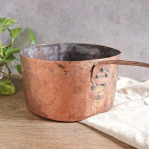 May include: A vintage copper pot with a handle and a riveted seam. The pot has a worn, aged patina and is slightly dented.