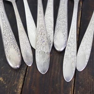 May include: Close-up of several silver spoons with a floral rose design. The spoons are arranged on a dark wooden surface. The spoons have a detailed pattern of roses and leaves on the handles and bowls.
