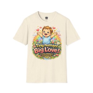 以下が含まれることがあります： クリーム色の半袖Tシャツ。腕を広げた笑顔の子供のグラフィックが描かれ、虹、花、ハートに囲まれています。テキストには「Tiny human! Big Love!」および「COLONNA STUDIO」とカラフルな文字で記されています。