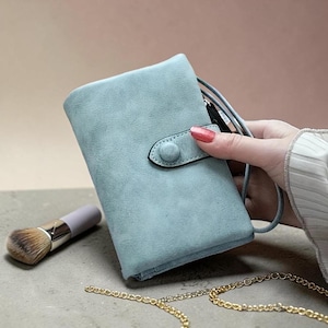 Op de afbeelding: Een lichtblauwe portemonnee met een drukknoopsluiting en een polsbandje. Een make-up kwast en een gouden ketting zijn ook zichtbaar. De portemonnee is gemaakt van een zacht, getextureerd materiaal.