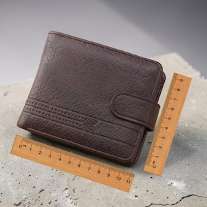Puede incluir: Una cartera de cuero marrón con una superficie texturizada y cierre de solapa. La cartera se muestra con dos reglas de madera, una que mide hasta 28 cm y la otra hasta 23 cm.