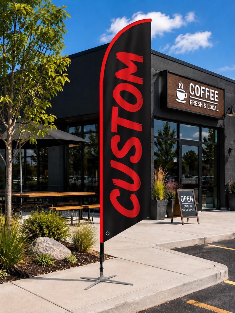 May include: A tall, black feather flag with a red border and the word CUSTOM printed in large, bold red letters. The flag is mounted on a silver metal cross-base stand, positioned upright on a concrete sidewalk outside a commercial building.