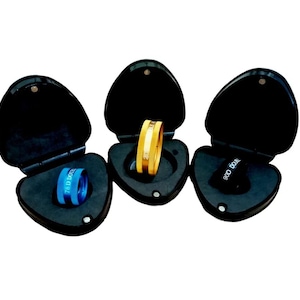 May include: Three black, heart-shaped cases, each open to reveal a ring or optical lens. One case holds a gold ring with a white stripe, another a blue lens with text, and the third a black lens with text.