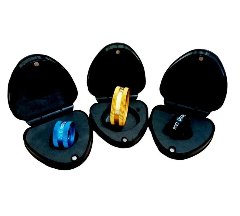 May include: Three black, heart-shaped cases, each open to reveal a ring or optical lens. One case holds a gold ring with a white stripe, another a blue lens with text, and the third a black lens with text.