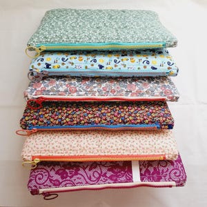 Peut inclure: Une pile de six pochettes rectangulaires en tissu matelassé, chacune présentant un motif distinct, notamment des imprimés floraux, géométriques et en dentelle. Chaque pochette est fermée par une tirette de fermeture éclair en plastique coloré.