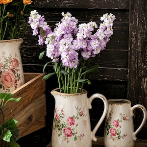 May include: A still life featuring floral arrangements in vintage-style pitchers. The pitchers are cream-colored with pink rose patterns and gold trim. One pitcher holds purple flowers, and another holds orange flowers. The scene is set on a wooden surface.