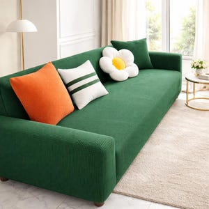 May include: A green sofa with a selection of cushions. The cushions include an orange square cushion, a white cushion with green stripes, a green square cushion, and a white flower-shaped cushion with a yellow centre. The sofa is in a bright room.