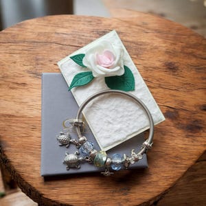 May include: A silver-toned charm bracelet featuring two turtle charms, several faceted glass beads in shades of blue and yellow, and metallic spacers. The bracelet rests on a grey box topped with a white paper card and a white and pink rose.