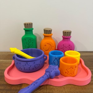 May include: A set of colourful silicone potion-making toys on a pink tray, including a purple cauldron, a purple wand, three bottles with wooden stoppers in green, orange, and pink, and three small cups in blue, orange, and yellow with embossed icons.