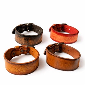May include: Four wide leather bracelets with metal buckles. One is black with a distressed texture, one is bright red with an orange gradient, one is light tan with a textured surface, and one is a medium brown with a smooth, polished finish.
