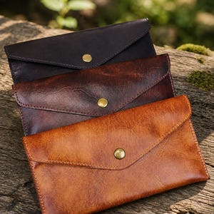 May include: Three rectangular envelope-style pouches in varying shades of brown leather. Each pouch features a pointed flap secured with a single brass-colored metal snap button. The colors range from a dark espresso to a medium reddish-brown.