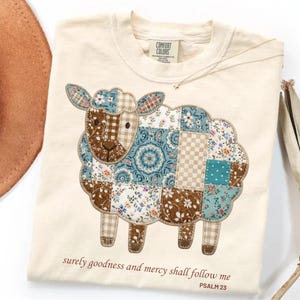 May include: A cream-colored t-shirt featuring a graphic of a sheep designed with a patchwork pattern of blue, brown, and white floral and gingham fabrics. The text "surely goodness and mercy shall follow me" and "PSALM 23" is printed in brown below.