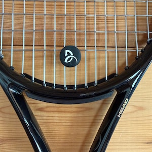 Könnte beinhalten: Ein schwarzer Tennisschläger mit silbernen Saiten und einem schwarzen Griff. Das Wort "HEAD" ist goldfarben auf der Seite des Schlägers aufgedruckt. Ein schwarzer Saitendämpfer mit weißem Logo ist in die Saiten eingesetzt.