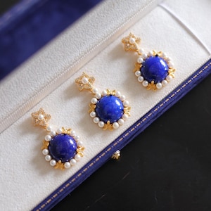 Może przedstawiać: Trzy złote kolczyki z głębokim, niebieskim, okrągłym kamieniem pośrodku, otoczone małymi białymi perłami. Kolczyki są prezentowane w białym pudełku z niebieskim wnętrzem i wykończeniem. Kolczyki mają kwiatowy wzór.
