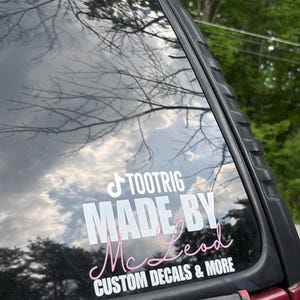 May include: A white and pink vinyl decal applied to a vehicle window. The decal features the text TOOTRIG, MADE BY, and CUSTOM DECALS & MORE in white, with the name McLead written in a pink cursive script font between the lines of white text.