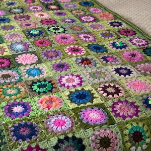 May include: A handmade crochet blanket featuring a floral pattern in a variety of colors. The blanket has a green background with squares of pink, purple, blue, and orange flowers. The blanket is approximately 60 inches by 80 inches.