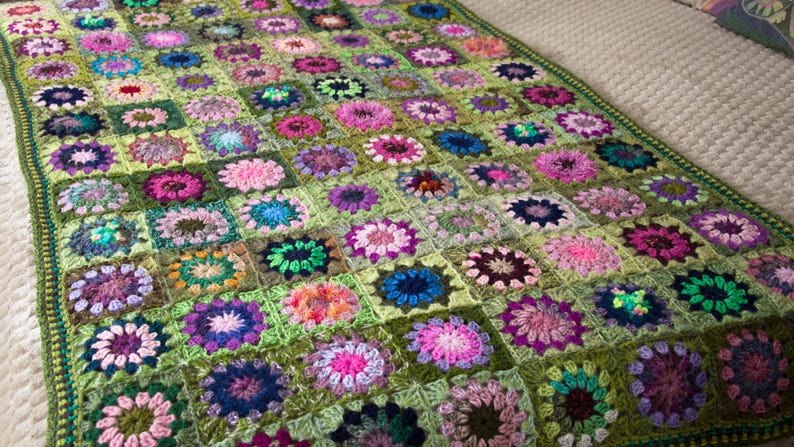 May include: A handmade crochet blanket featuring a floral pattern in a variety of colors. The blanket has a green background with squares of pink, purple, blue, and orange flowers. The blanket is approximately 60 inches by 80 inches.