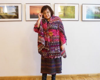 May include: A woman wearing a colorful crocheted shawl with a large button in the center. The shawl is made of various shades of pink, purple, and brown yarn. The woman is standing in front of a white wall with framed pictures.