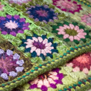 May include: A close-up of a handmade crochet blanket. The blanket features a repeating pattern of colorful floral squares in shades of green, purple, pink, and orange. The edges are finished with a green border.