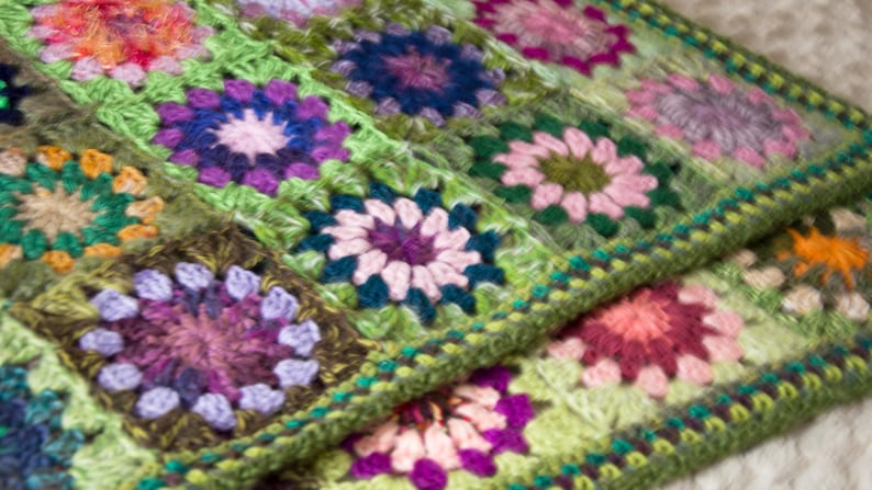 May include: A close-up of a handmade crochet blanket. The blanket features a repeating pattern of colorful floral squares in shades of green, purple, pink, and orange. The edges are finished with a green border.