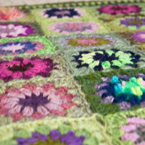 May include: A handmade crochet blanket featuring a floral pattern. The blanket is composed of numerous squares, each showcasing a flower design in shades of pink, purple, green, and blue. The overall color palette is soft and inviting, with a focus on texture.