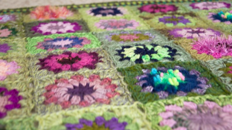 May include: A handmade crochet blanket featuring a floral pattern. The blanket is composed of numerous squares, each showcasing a flower design in shades of pink, purple, green, and blue. The overall color palette is soft and inviting, with a focus on texture.