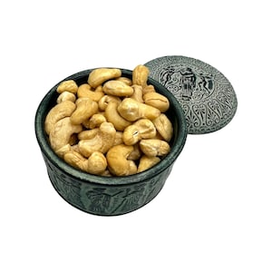 May include: A round, dark green container filled with light brown cashew nuts. The container and its matching lid feature embossed figures and Greek text. The lid is placed beside the container, displaying intricate patterns and a central figure.
