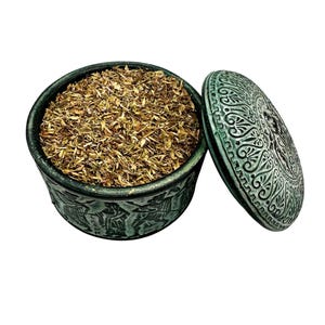 May include: A teal ceramic container with a lid, filled with dried herbs. The container has an embossed design around the sides and on the lid. The lid is partially open, revealing the contents.