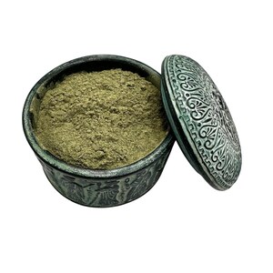 Pode incluir: Um recipiente redondo verde-escuro com tampa a condizer, apresentando figuras em relevo na lateral e padrões intrincados na tampa. O recipiente está cheio de um pó fino castanho-claro, exibindo um acabamento texturizado e envelhecido.