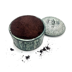 May include: A round, teal-colored metal container filled with dark brown, granular powder. The exterior of the container and its separate, circular lid feature embossed, classical-style figures and intricate decorative patterns.