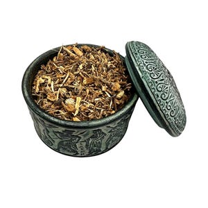 Può includere: Un barattolo rotondo in ceramica grigio scuro con coperchio abbinato, entrambi caratterizzati da intricati rilievi di figure e motivi. Il barattolo è riempito con una miscela di erbe legnose essiccate marrone chiaro e piccoli frammenti di radici.
