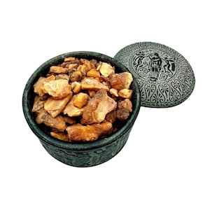 May include: A dark green container filled with light brown and tan resin pieces. The container has a matching lid with a detailed relief design. The lid is partially open, revealing the contents.