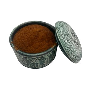 May include: A round, dark gray container with intricate embossed figures and patterns on the exterior, filled with a fine, reddish-brown powder. The matching circular lid, featuring detailed scrollwork and carvings, rests beside the open container.