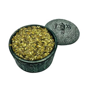 May include: A dark green, round container filled with dried, light green leaves, with a matching lid. The lid features a detailed, grey, circular design with figures and patterns. The container and lid appear to be made of a stone-like material.