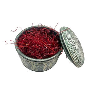 May include: A round, dark grey ceramic container with intricate embossed patterns on the exterior and lid, filled with a generous amount of vibrant red saffron threads. The lid is placed beside the open container, revealing the textured interior.