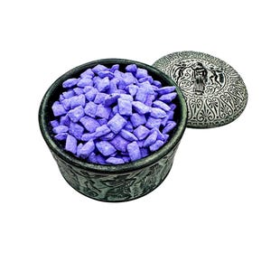 May include: A decorative, dark gray metal bowl filled with small, square, lavender-colored items. The bowl has an embossed design around the exterior. A matching lid with an ornate design is next to the bowl.