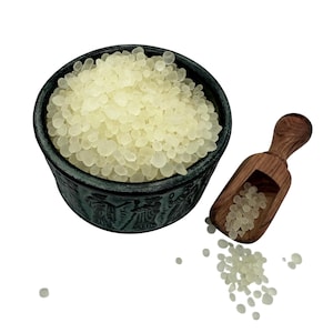 Peut inclure: Un bol vert foncé avec des figures en relief rempli de petites perles de résine translucides jaune pâle. Une petite pelle en bois avec un motif de grain visible repose à côté du bol, contenant quelques perles, avec d'autres éparpillées.