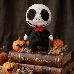 Op de afbeelding: Gehaakte Jack Skellington-pop met een witte kop, zwarte ogen en een gestikte glimlach. De pop draagt een zwart pak met een rode vlinderdas. Het wordt gepresenteerd met oranje pompoenen op antieke boeken.