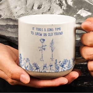 Pode incluir: Caneca de cerâmica creme com base e alça marrons. A caneca apresenta ilustrações florais azuis e o texto "IT TAKES A LONG TIME TO GROW AN OLD FRIEND." Os nomes "Diana" e "Anna" também estão impressos.