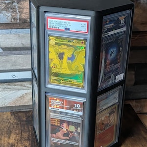May include: A black hexagonal rotating display stand featuring multiple vertical slots for graded trading cards. The display holds several plastic-encased cards, including a gold Mega Charizard X and a Giovanni's Charisma card, each with visible labels.