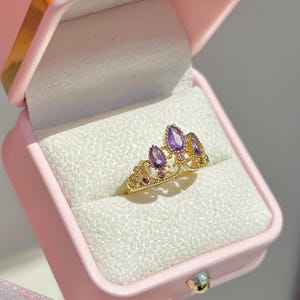 May include: A gold-coloured crown ring featuring purple teardrop-shaped gemstones and smaller round purple gems. The ring is presented in a pink jewellery box with a white interior.