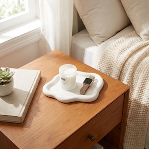 May include: A white, organically shaped tray holds a lit candle and a smartwatch with a tan band. The tray sits on a wooden nightstand next to a closed book and a small potted succulent. The scene is bathed in soft, natural light.