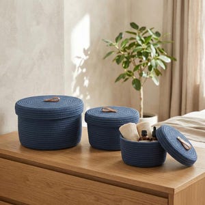 May include: Three round, navy blue rope storage baskets with matching lids. Each lid features a small, brown leather pull tab. The smallest basket is open, revealing two beige fabric cloths and two small glass bottles with dark droppers.
