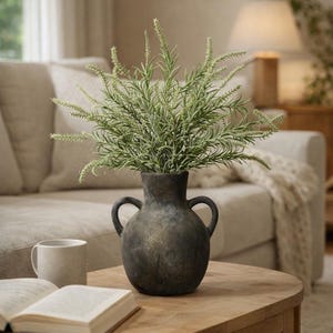 May include: A dark gray, textured vase with two handles holds a lush arrangement of green and white faux rosemary. The vase sits on a wooden table, with a mug and open book nearby, creating a cozy, home decor scene.
