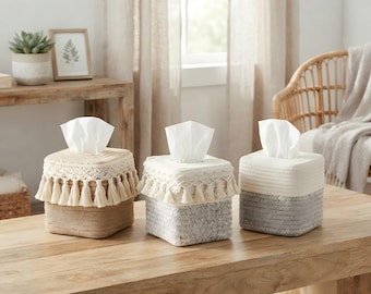 May include: Three square tissue box covers made of woven rope. The first is tan with a cream macrame fringe. The second is a grey and white woven pattern with a cream macrame fringe. The third is a solid cream woven rope design.