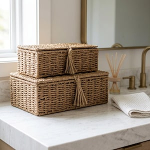 May include: Two rectangular woven storage boxes made of natural tan fiber, stacked one on top of the other. Each box features a matching lid secured with a braided rope loop and a decorative hanging tassel centered on the front edge.
