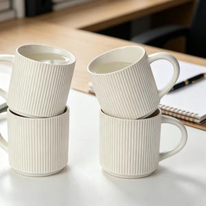 May include: Four ivory-colored ceramic mugs stacked on a white surface. Each mug has a vertical ribbed design and a curved handle. The mugs are filled with a clear liquid, possibly water.