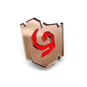 Op de afbeelding: Een rode en gouden emaille pin met een gestileerd swirl-ontwerp. De pin is gevormd als een schild.