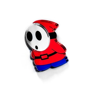 May include: An acrylic pin featuring a cartoon character with a white mask, black eyes, and a red hooded cloak. The character is wearing blue trousers.