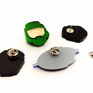 Pode incluir: Uma cole&ccedil;&atilde;o de alfinetes de acr&iacute;lico em v&aacute;rias formas e cores. Um alfinete &eacute; um capacete verde e dourado, outros s&atilde;o pretos, cinzentos e azuis. Cada alfinete tem um verso prateado. O logotipo Chinook Crafts est&aacute; no canto inferior esquerdo.